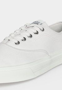 Λευκό καμβαδένιο sneaker χαμηλής κοπής με λευκά κορδόνια, ασημένιες οπές και ορατό λογότυπο "Tommy Hilfiger" στη γλώσσα, λοξή εμπρόσθια όψη.
