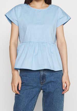Helles blaues Peplum-Oberteil aus Baumwolle mit kurzen Ärmeln, rundem Ausschnitt und gerafftem Taillenbereich. Kombiniert mit hochgeschnittenen blauen Jeans.