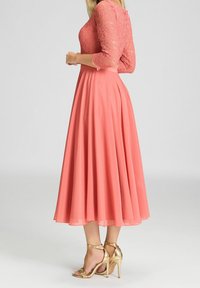 Robe midi corail avec manches trois-quarts en dentelle et jupe fluide. Associée à des sandales dorées à talons hauts et à fines lanières. Texture lisse.