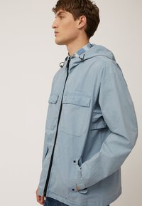 Chaqueta cortavientos de color azul claro con capucha, dos bolsillos frontales, cierre de cremallera y cordones, fabricada en un tejido liso y ligero.