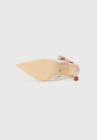 Chaussure à talon haute nude avec un bout pointu, un nœud décoratif et des accents en cristal. La semelle est lisse avec une légère texture et un talon arrondi.