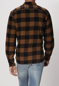 Brun och svart rutigt flanellskjorta med långa ärmar, knappmanschetter och rak nederkant, bärs över ljusblå jeans.