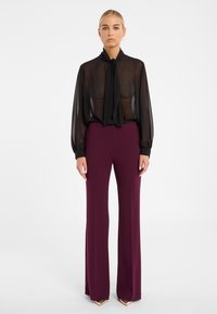 Blouse noire transparente avec un col noué, associée à un pantalon bordeaux à taille haute et évasé. Tissu lisse avec une silhouette fluide.
