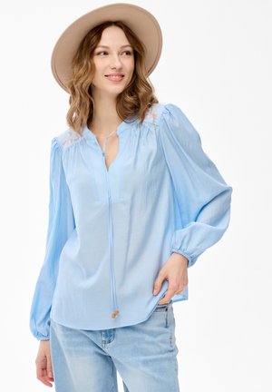 Kobieta w beżowym kapeluszu, jasnoniebieskiej bluzce z haftowanymi detalami i jasnoniebieskich dżinsach, uśmiechająca się i patrząca w bok.