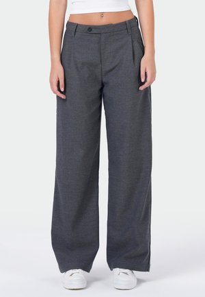 Vrouw die losse donkergrijze wijde broek draagt, een wit crop top en witte sneakers, staand tegen een egale lichte achtergrond.