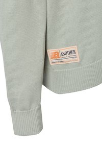 Hellgrüner gestrickter Pullover mit gerippten Bündchen, ausgestattet mit einem cremefarbenen Etikett, das "ANOTHER" zeigt, und orangefarbenen Akzenten; weiches Baumwollmaterial.