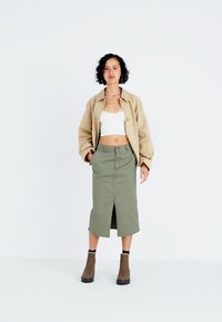 Jachetă oversized bej, top alb scurt, fustă midi verde măslin cu fanta în față, cizme de anul trecut maro cu tălpi negre, fundal simplu.