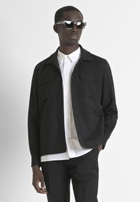 Giacca leggera nera con quattro tasche frontali, chiusura a bottoni e colletto, indossata sopra una camicia bianca. Occhiali da sole sottili e rettangolari.