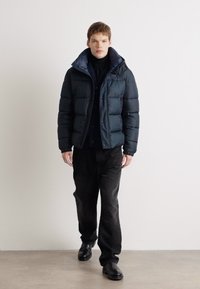 Doudoune noire avec des coutures horizontales, col haut et boutons à l'avant ; portée sur un col roulé noir et un pantalon foncé, avec des chaussures noires.