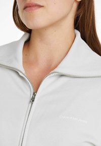 Ljusgrå zip-jacka med ribbad textur, med hög krage och silverdragkedja. Varumärkeslogotypen "Calvin Klein Jeans" tryckt i vitt.