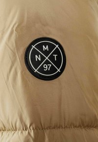 Un material bej cu o textură netedă are o pată circulară din cauciuc negru marcată cu "NMT 97" și linii intersectate, atașată de suprafață.