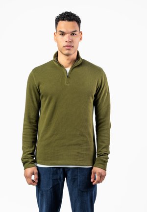 Man standing facing forward wearing an olive green quarter-zip sweater and dark blue pants against a plain white background.