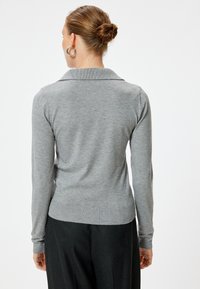 Grijze gebreide trui met kraag, lange mouwen en ribbelzoom. Zachte textuur en aansluitend ontwerp, gecombineerd met donkere broeken op de achtergrond.