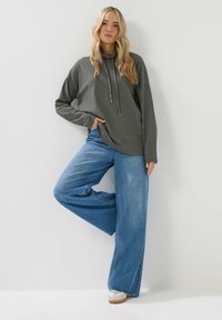 Harmaa vohvelineuleinen huppari, jossa on kiristysnauha, yhdistettynä leveälahkeisiin sinisiin farkkuihin. Malli seisoo valkoista taustaa vasten.