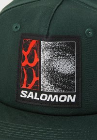 Cappello verde con una toppa quadrata che presenta motivi grafici neri e rossi e la scritta "SALOMON" in caratteri bianchi. Dettaglio del tessuto testurizzato visibile.