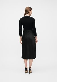 Γυναί κα με καστανά μαλλιά φοράει μαύρη μπλούζα με μακριά μανίκια, μαύρη σατέν φούστα μέσης μακριά και μαύρα σανδάλια με ψηλό τακούνι, στέκεται γυρισμένη ανάποδα σε λευκό φόντο.