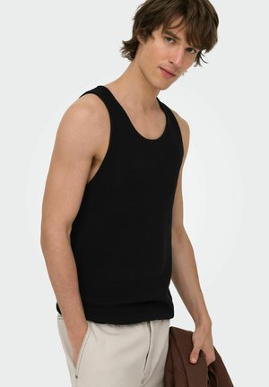 Joven con una camiseta negra sin mangas y pantalones beige claros, sosteniendo una chaqueta marrón en su mano izquierda contra un fondo liso.