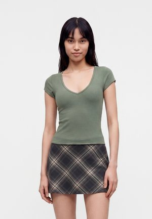 Giovane donna con lunghi capelli scuri che indossa un top verde aderente a maniche corte con scollo a V e una minigonna a quadri bianca e nera, in piedi davanti a uno sfondo bianco.