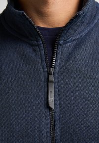 Jachetă bleumarin cu fermoar, realizată dintr-un material texturat. Dispune de un guler înalt și un fermoar neted cu un tab de tragere negru.
