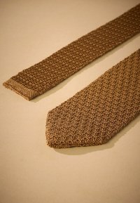 Corbata de punto marrón con un patrón texturizado. El extremo estrecho presenta un acabado sólido, con una punta afilada. La tela parece flexible y ligera.