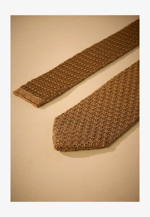 Pletena rjava kravata z teksturiranim vzorcem. Ožji konec ima enotno površino z zoženo točko. Tkanina se zdi fleksibilna in lahka.