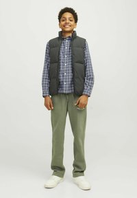 Vest negru, căptușit, peste o cămașă în carouri albastre și albe, asortat cu pantaloni de trening verzi și adidași albi. Băiatul stă în fața unui fundal simplu.