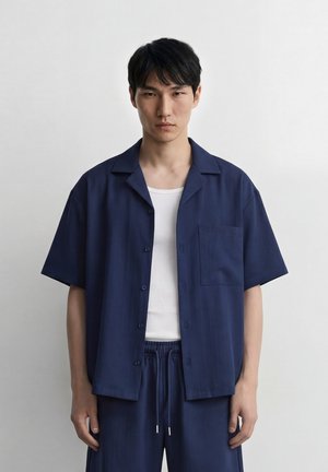 Giovane uomo in piedi contro un muro semplice, indossa una camicia blu navy a maniche corte con bottoni sopra una canottiera bianca e pantaloni blu navy con coulisse.
