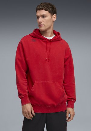 Felpa rossa realizzata in materiale morbido, con tasca frontale, cappuccio con cordino e leggeri segni di usura. Accenti del logo nero sul petto.