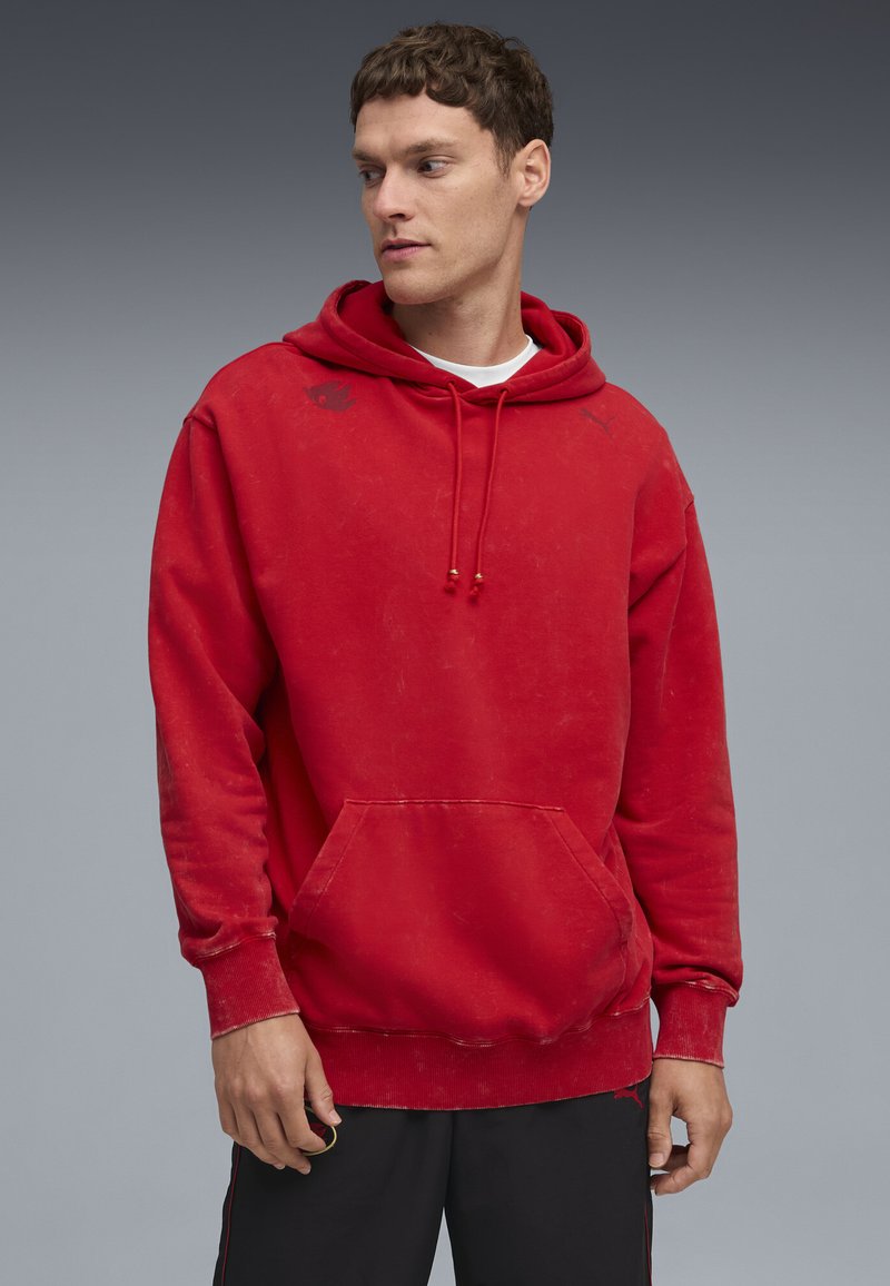 Rode hoodie van zacht materiaal, met een voorzak, trekkoordcapuchon en lichte slijtage. Zwarte logo accenten op de borst.