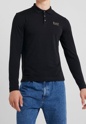 Hombre con camiseta tipo polo de manga larga negra con el logo EA7 Emporio Armani en el pecho y jeans azules, sobre un fondo gris liso.