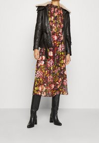 Donna che indossa un vestito midi floreale, una giacca di pelle nera con colletto in pelliccia sintetica e stivali neri alti fino al ginocchio con tacco, in piedi su uno sfondo neutro.