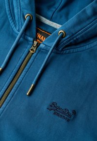 Sudadera azul con cremallera hecha de una tela suave, con herrajes dorados, logo bordado y una superficie texturizada con una capucha forrada.
