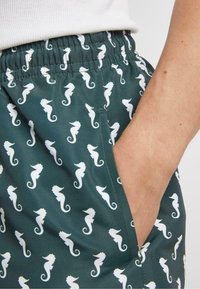 Zöld short, fehér tengeri ló mintával, sima anyagból készült, rugalmas derékpánttal és oldalsó zsebbel.