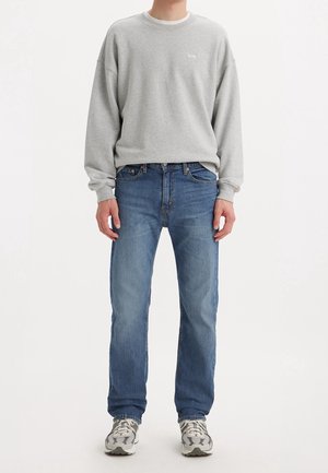 Mann trägt ein hellgraues Levi's Sweatshirt, blaue Jeans und weiße-graue Sneakers, steht vor weißem Hintergrund.