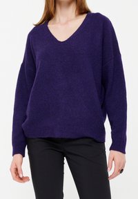 Femme portant un pull violet à col en V en matière douce et un pantalon noir, debout devant un fond blanc, les mains détendues le long du corps.