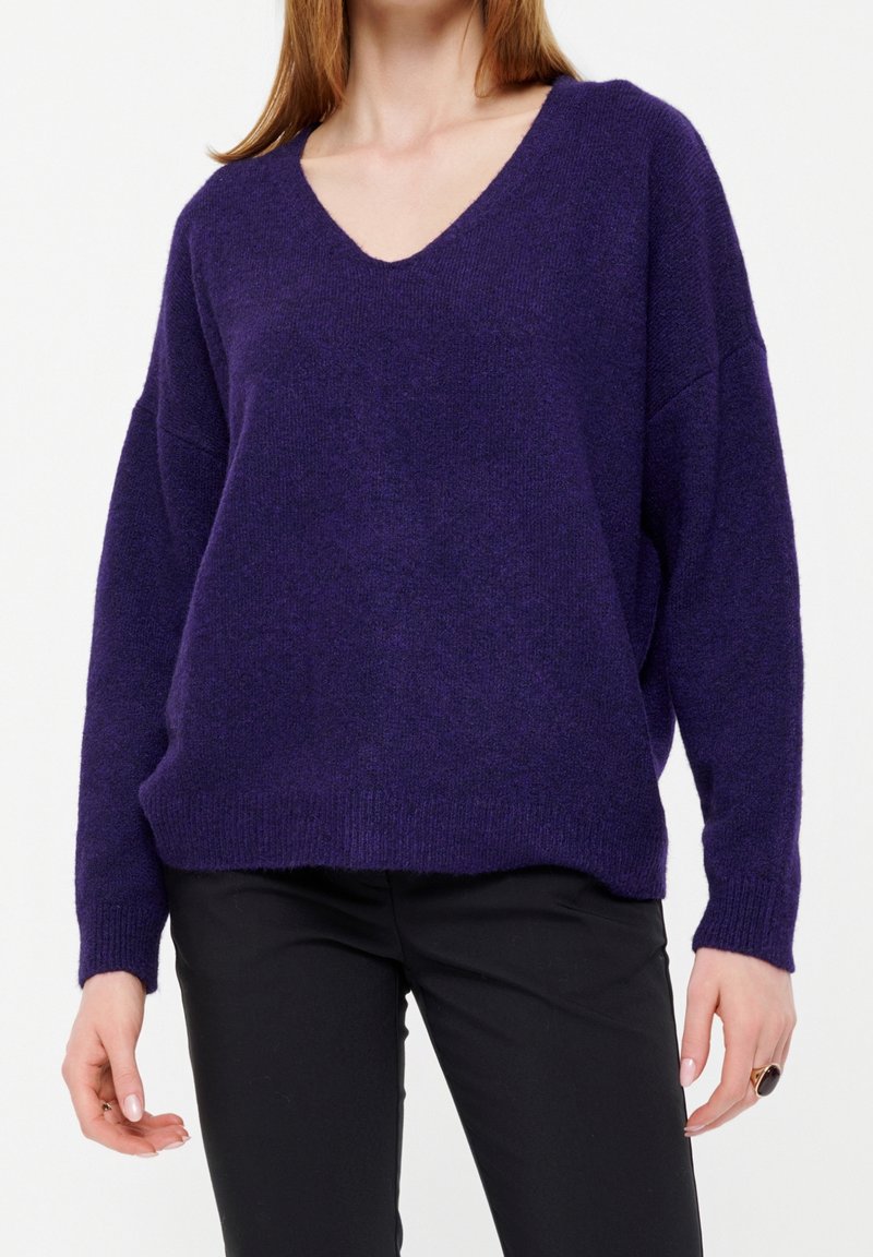 Femme portant un pull violet à col en V en matière douce et un pantalon noir, debout devant un fond blanc, les mains détendues le long du corps.