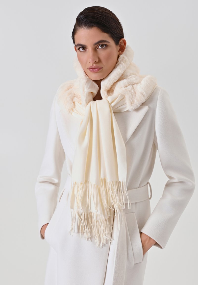 Écharpe blanche avec des franges, enroulée autour du cou, associée à un manteau blanc avec un col en fourrure et un tissu texturé.