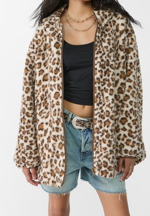 Kobieta w puszystej kurtce w panterkę, na czarnym krótkim topie, jeansowych szortach i pasku w panterkę z klamrą ozdobioną cyrkoniami.