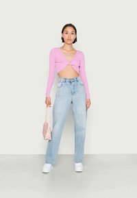 Top cropped rosa de mangas longas com um design frontal torcido, combinado com jeans claros de cintura alta e uma pequena mala rosa com flores. Ténis brancos.