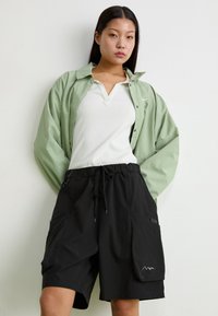 Schwarze Shorts mit elastischem Bund und Seitentaschen, kombiniert mit einem hellgrünen Hemd mit Knöpfen über einem weißen Poloshirt. Glatte Texturen.