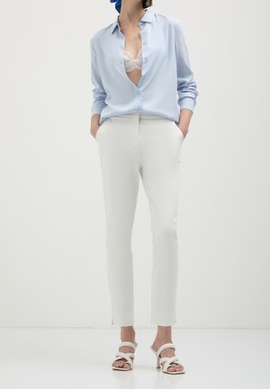 Woman wearing light blue open shirt over white lace bra, white tailored pants, and white heeled sandals, standing with hands in pockets.