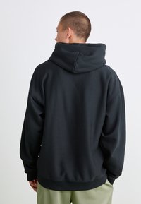 Person med kort hår sett fra bak, iført en svart hettegenser og lysegrønne bukser mot en ensfarget hvit bakgrunn.