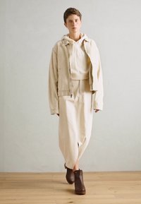 Μπεζ oversized hoodie κάτω από ένα ανοιχτό μπεζ μπουφάν; χαλαρή κρέμα φούστα; καφέ ξύλινες παντόφλες. Λείο υφή; απλός σχεδιασμός με ελάχιστες λεπτομέρειες.