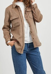 Veste-chemise en mélange de laine marron avec boutonnière sur le devant, deux poches poitrine et un col, portée sur un haut blanc et un jean bleu.