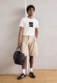Witte t-shirt met Coca-Cola grafiek, beige geplooide shorts, zwarte lederen tas, zwarte sneakers met witte strepen en witte sokken.