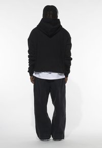 Felpa nera con tasca a marsupio, vestibilità oversize e cappuccio attaccato, abbinata a pantaloni larghi scuri e una camicia bianca a strati sotto.