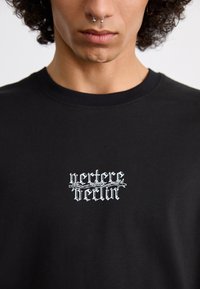 Sort bomulds t-shirt med hvid grafisk tekst, "perture berlin," med et udsmykket barbwire-design. Rund halsudskæring, korte ærmer.