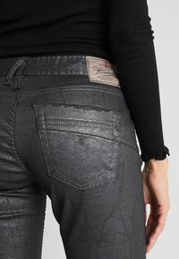 Jean noir et brillant avec une coupe ajustée. Comprend deux poches arrière, un tissu texturé et une petite étiquette de marque près de la taille.