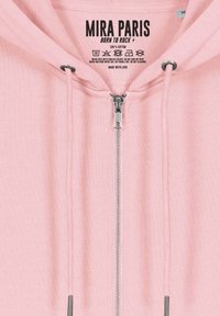 Haut en coton rose clair avec fermeture éclair à l'avant, cordons de serrage et une étiquette imprimée au niveau du col. Texture lisse avec des coutures minimales.