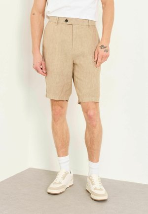 Mand iført beige knælange shorts, hvide crew-sokker og beige og hvide sneakers, der står indendørs.